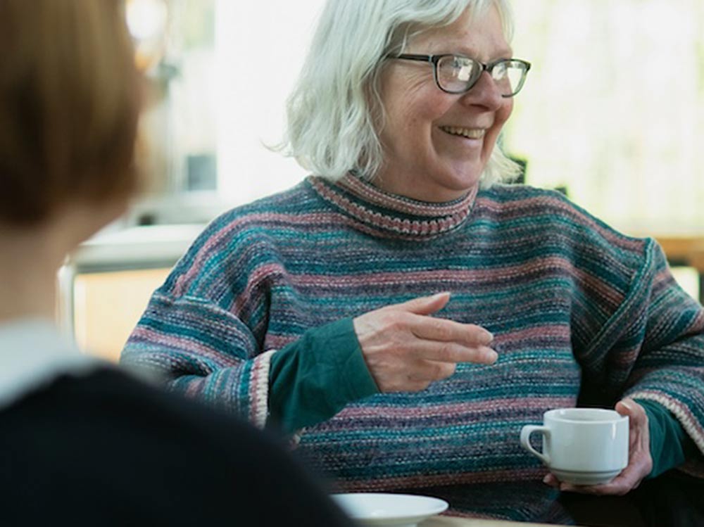 An older woman holding a coffee cup talks to friends in a cafe